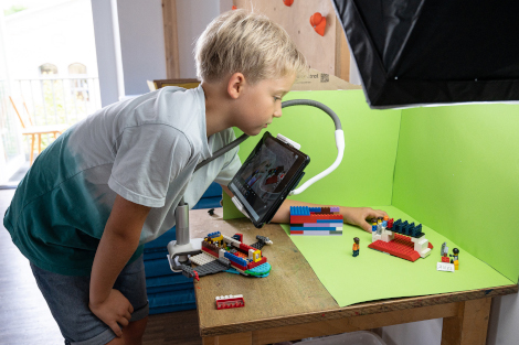 Auf einem Tisch vor einem grünem Hintergrund ist eine Szene aus Lego aufgebaut; davor ist ein Tablet installiert, vor das sich ein Junge beugt.