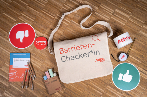 Eine Tasche mit Aufdruck 'Barrieren-Checker*in', daneben Straßenkreiden, Rolle mit Absperrband, Notizheft und Aufkleber