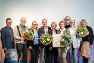 Neun Menschen stehen nebeneinander auf einer Bühne. Einige halten einen Blumenstrauß, eine Frau hält eine gerahmte Urkunde.