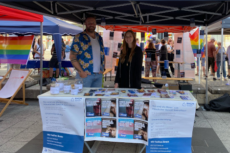 Ein Mann links und eine Frau rechts stehen unter einem Pavillon an einem Infostand, es gibt einige Regenbogendekorationen.