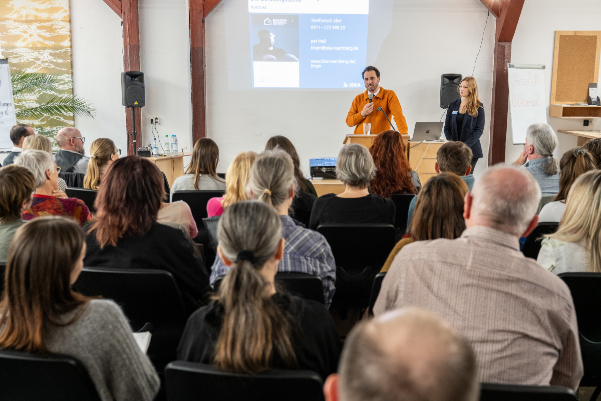 Ein Mann steht vor einem Publikum und h&auml;lt einen Vortrag. Eine Frau, die Moderatorin, steht neben ihm.