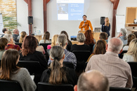 Ein Mann steht vor einem Publikum und hält einen Vortrag. Eine Frau, die Moderatorin, steht neben ihm.