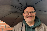 Ein Mann mit Schnurrbart hält einen Schirm und grinst in die Kamera