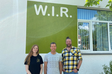 Eine junge Frau und zwei junge Männer stehen draußen nebeneinander vor einer Hauswand, auf der vor grünem Hintergrund in weißen Buchstaben steht W.I.R.