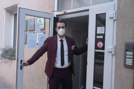 Ein Mann mit dunkelrotem Anzug hält eine Eingangstür einladend offen.
