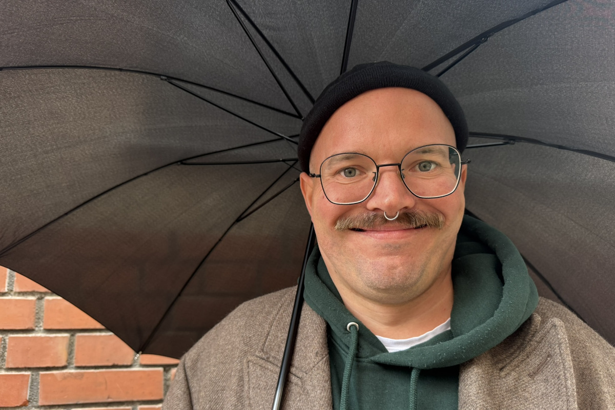 Ein Mann mit Schnurrbart steht draußen mit Regenschirm und grinst in die Kamera.