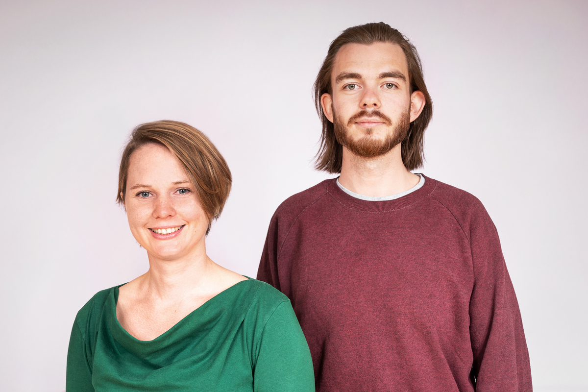 Eine junge Frau mit blonden Harren und grünes Shirt und ein junger Mann mit Bart und rotem Pullover blicken freundlich in die Kamera.