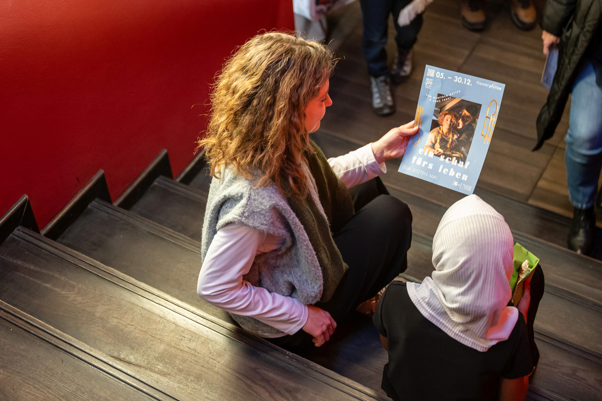 Eine Frau und ein Mädchen sitzen auf den Treppenstufen vor einem Theatersaal und schauen gemeinsam auf das Programmheft des Theaterstücks, das sie gleich anschauen werden.