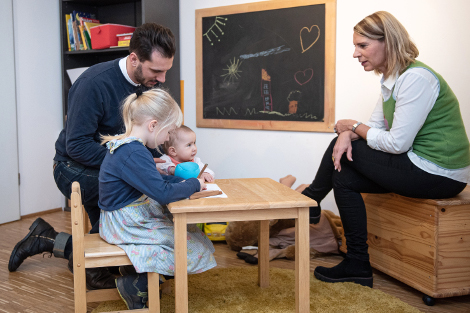 Ein Mann hockt bei zwei kleinen, malenden Kindern am einen niedrigen Tisch. gegenüber sitzt eine Frau am Tisch und lacht.