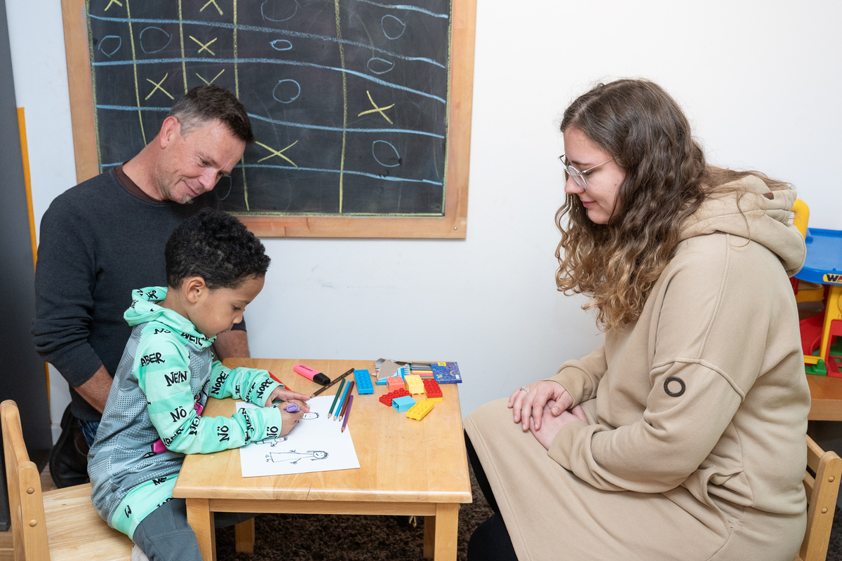 Ein Vater sitzt mit seinem fünfjährigen Sohn, der etwas malt, an einem niedrigen Tisch. Gegenüber sitzt eine Ehrenamtliche am Tisch und lächelt.