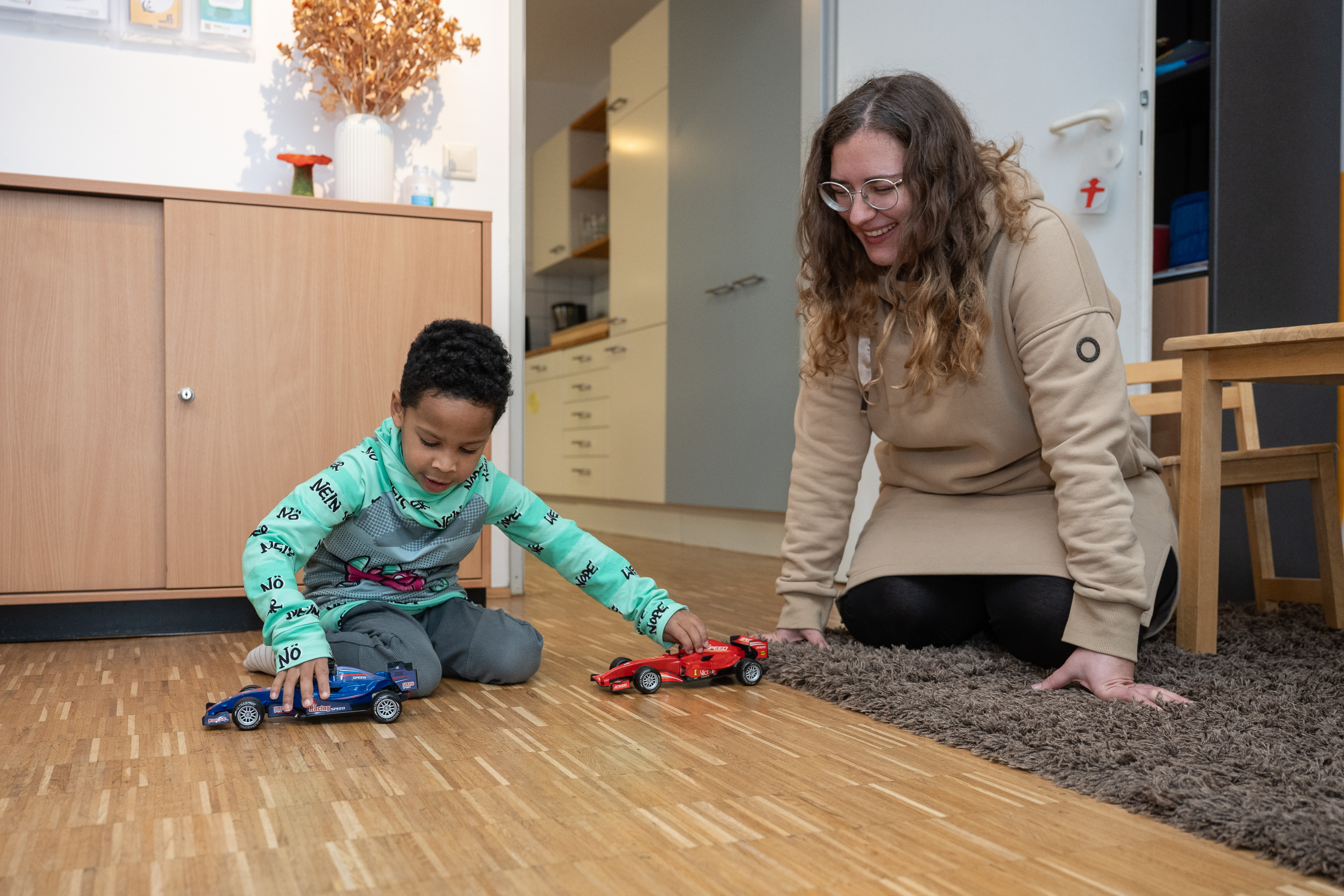 Ein fünfjähriger Junge sitzt auf dem Boden und spielt mit Autos. Neben ihm sitzt eine Ehrenamtliche und beobachtet ihn lächelnd.