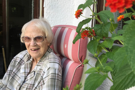 Eine weißhaarige, schlanke Frau sitzt in einem Gartenstuhl neben einer blühenden Balkonpflanze und lacht in die Kamera.