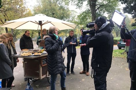 Das Caféfahrrad und Ehrenamtliche stehen links im Bild, davor eine ARD-Reporterin mit Mikro, rechts ein Kameramann, der sie filmt.