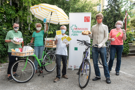 Fünf Menschen mit Mund-Nasen-Schutzmasken stehen neben Fahrrädern. Sie halten ein Tablett mit Muffins und verpackte Mund-Nasen-Schutze hoch.