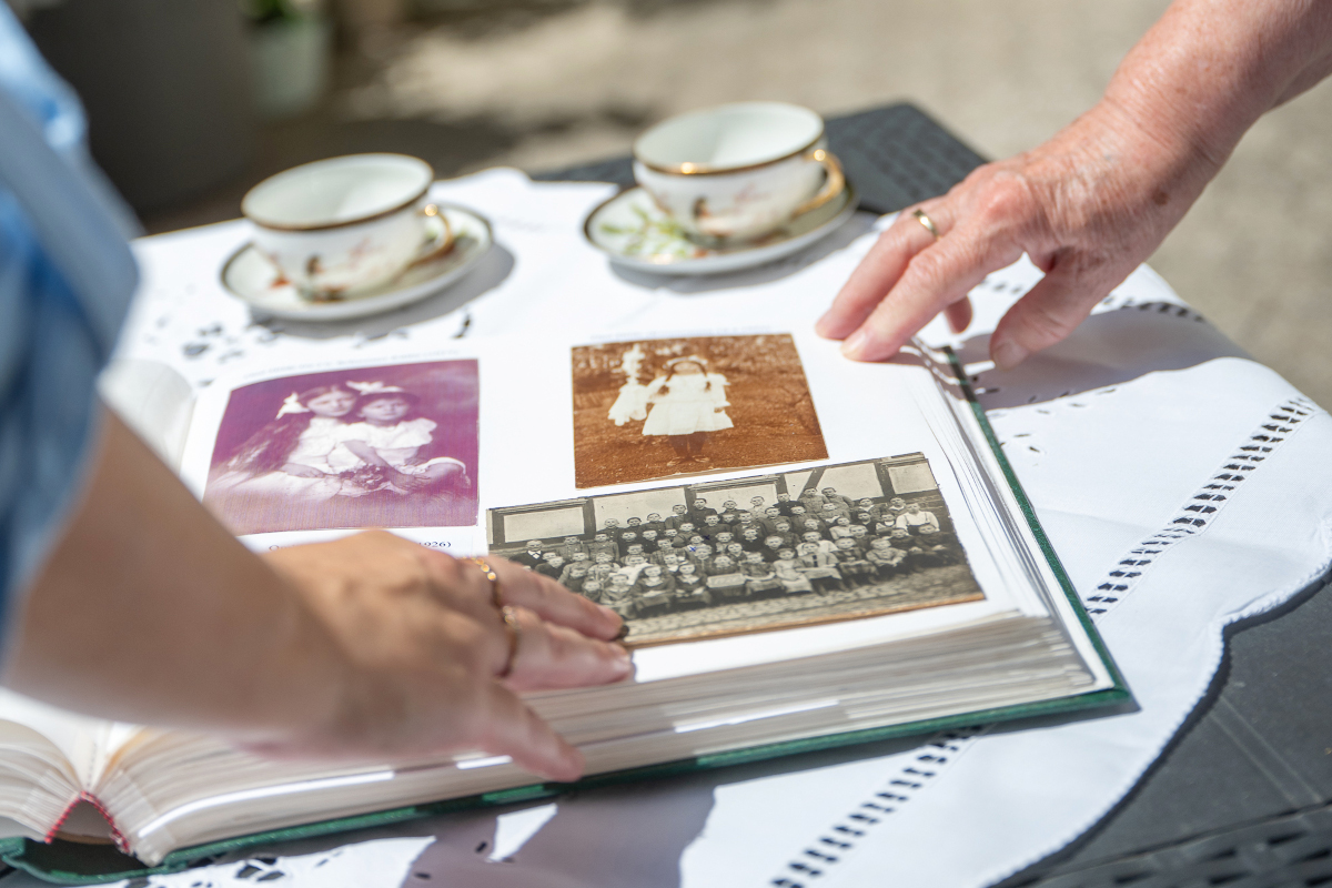Eine alte und eine jüngere Hand ruhen auf einem aufgeschlagenen Fotoalbum mit alten Schwarz-weiß-Aufnahmen, das auf einem Kaffeetisch im Freien liegt. Auf dem Tisch stehen zwei Kaffeetassen eines Services aus den 1940ern.