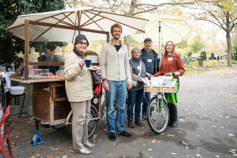 Vor einem umgebauten Lastenfahrrad mit Kuchentresen und Sonnenschirm stehen mehrere Personen, u.a. mit Kaffeetasse in der Hand