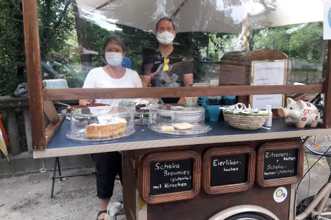 Hinter einer Kuchentheke auf einem Fahrradaufbau stehen zwei Personen mit Maske