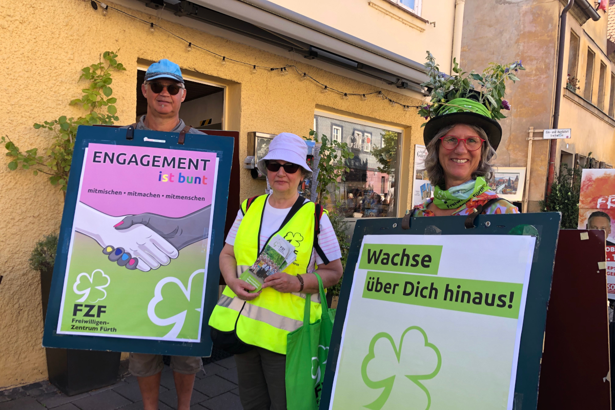 Ein Mann und zwei Frauen stehen im Freien vor einem Haus. Der Mann und eine der Frauen tragen Plakate am Körper, die zweite Frau hält Flyer in der Hand.