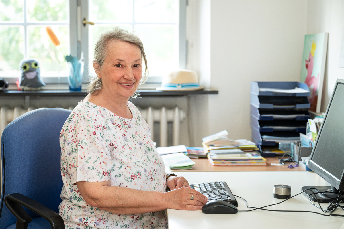 Eine Frau mit bunter Bluse sitzt an einem Schreibtisch vorm PC und lacht in die Kamera.