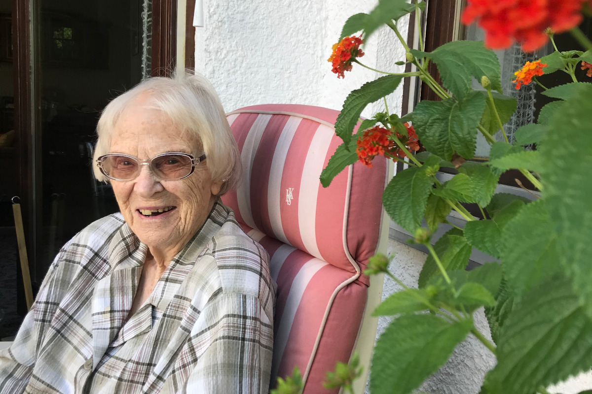 Eine weißhaarige, schlanke Frau sitzt in einem Gartenstuhl neben einer blühenden Balkonpflanze und lacht in die Kamera.