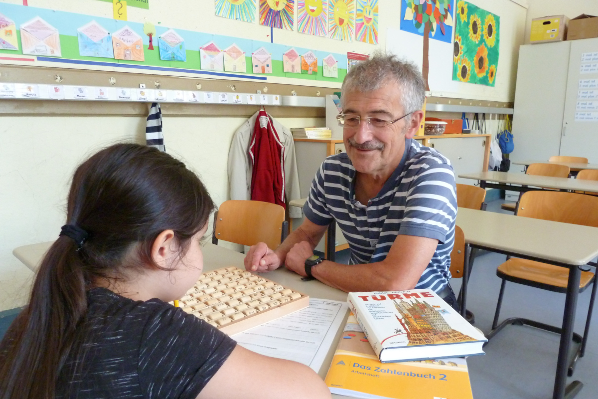 Ein Mann sitzt an einem Schreibtisch in einem Klassenzimmer. Ihm gegenüber eine Schülerin, gebeugt über Unterrichtsmaterial.