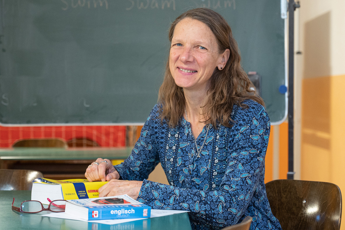 Eine Frau mit blauer Bluse und langen Haaren sitzt fröhlich lachend an einem Schreibtisch, vor ihr liegt ein Englischwörterbuch