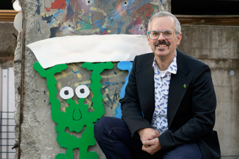 Ein Mann mit offenem Jacket und Brille hockt lächelnd vor einer bunten Betonwand, hinter ihm ist die Figur eines lustigen, grünen Monsters zu sehen.