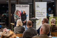 Zwei Frauen stehen vor einem Publikum, die eine spricht gerade mit Mikrofon in der Hand, die andere grinst in sich hinein.