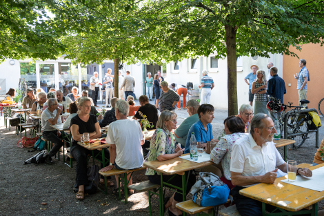 An Biertischen unter Bäumen sitzen viele Menschen im Sonnenschein vor kalten Getränken und unterhalten sich angeregt.