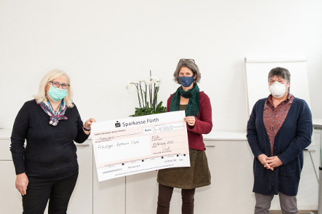 Drei Frauen mit Mundschutz stehen mit Abstand zueinander vor einem weißen Hintergrund und halten gemeinsam einen Check.