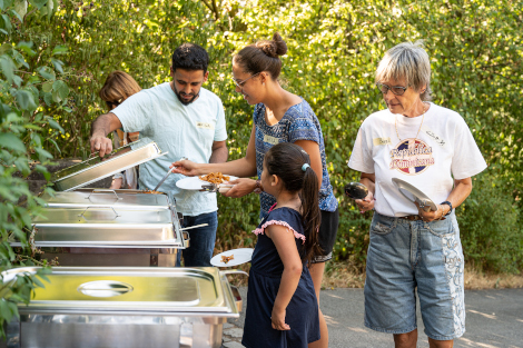 An einem Buffet draußen im Sonnenschein stehen mehrere Personen, u.a. ein kleines Mädchen und holen sich Essen.