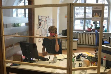 Ein junger Mann mit Mund-Nasen-Schutzmaske sitzt hinter einer Plexiglasscheibe an einem Schreibtisch und telefoniert.
