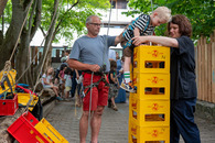 in kleiner Junge klettert mit Seilen gesichert an aufeinandergestapelten Getränkekästen empor. Eine Frau und eine Mann helfen ihm dabei.