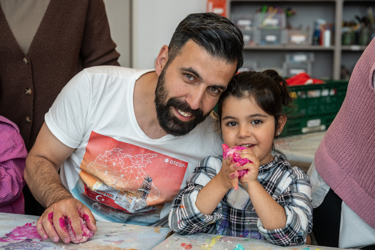 Ein Vater und seine Tochter, arabischer Herkunft, sitzen am Werktisch, haben Modelliermasse in der Hand und lächeln in die Kamera.