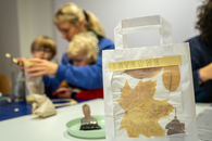 Eine Mutter sitzt mit zwei Kindern am Tisch und bastelt eine Laterne. Im Vordergrund steht eine bereits fertiggestellte Laterne.