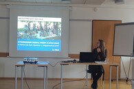 Eine Frau sitzt vor einer Leinwand, auf die eine Präsentation projiziert wird, und blickt auf diese.