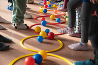 Ein aufgebautes Bewegungsspiel mit Ringen und bunten kleinen Bällen auf dem Boden. Die Kamera ist auf den Boden gerichtet, links und rechts von den Ringen stehen Kinder, deren Füße man sieht.