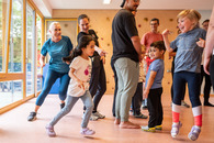Kinder und Erwachsene stehen in bzw. rennen durch eine Turnhalle. Ein Kind springt vor Freude in die Luft.