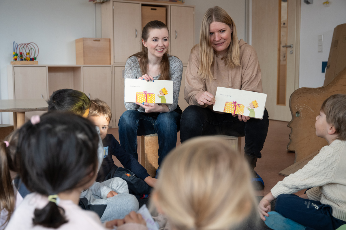 Zwei Frauen sitzen vor einer Gruppe von Kindergartenkindern und halten Bilderbücher hoch, aus denen sie gemeinsam vorlesen.