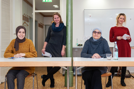 Im Vordergrund sitzen zwei muslimische Frauen mit Hijab an einem Tisch, im Hintergrund stehen zwei Frauen vom ISKA, alle lächeln in die Kamera.