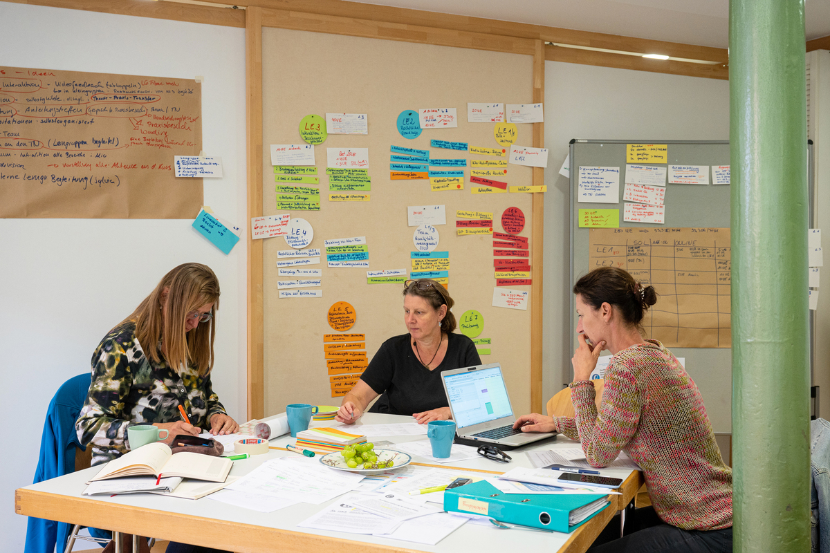 An eine Tisch mit vielen Arbeitmaterialien sitzen drei Frauen im Gespräch, im Hintergrund eine Pinnwand mit vielen Notizen