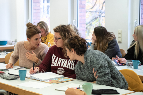 Drei Frauen sitzen nebeneinander an einem Tisch, vor ihnen liegen Arbeitsmaterialien, sie beugen sich zu einander und diskutieren.