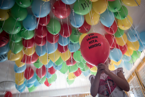 Unter der Zimmerdecke schweben ganz viele bunte Luftballons, ein Mädchen steht vorn und hält einen Luftballon mit der Aufschrift: 10 JAHRE KITA MIKRO