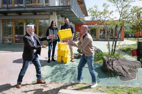 Ein Mann wirft einem anderen eine große gelbe Box zu. Sie stehen draußen vor einem gebäude, im Hintergrund schauen zwei Erwachsene zu.