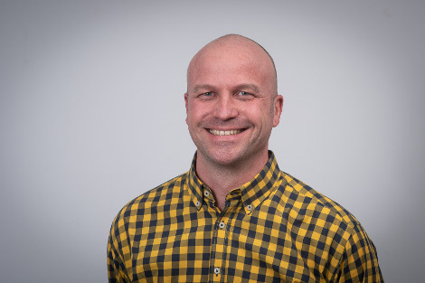 Porträtfoto: Ein Mann mit gelbschwarz-kariertem Hemd lächelt freundlich in die Kamera
