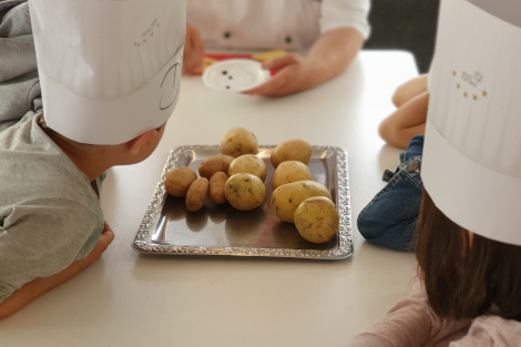 Auf einem Tisch liegen Kartoffeln, mehrere Kinder mit weißen Kochmützen sitzen um den Tisch und betrachten sie.
