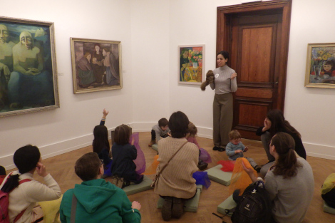 Kinder sitzen am Boden in einem großen Raum mit Bildern an den Wänden, vor ihnen steht eine Frau mit einer Handpuppe.