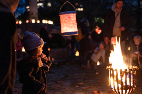 Ein kleines Mädchen schaut auf eine Feuerschale, drumherum sieht man Laternen und andere Menschen.