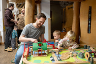 Ein Vater spielt mit zwei kleinen Kindern mit einer Holzeisenbahn in der Kita. Im Hintergrund stehen zwei weitere Väter, die sich unterhalten. Ein Junge schmiegt sich an einen dieser beiden Väter.