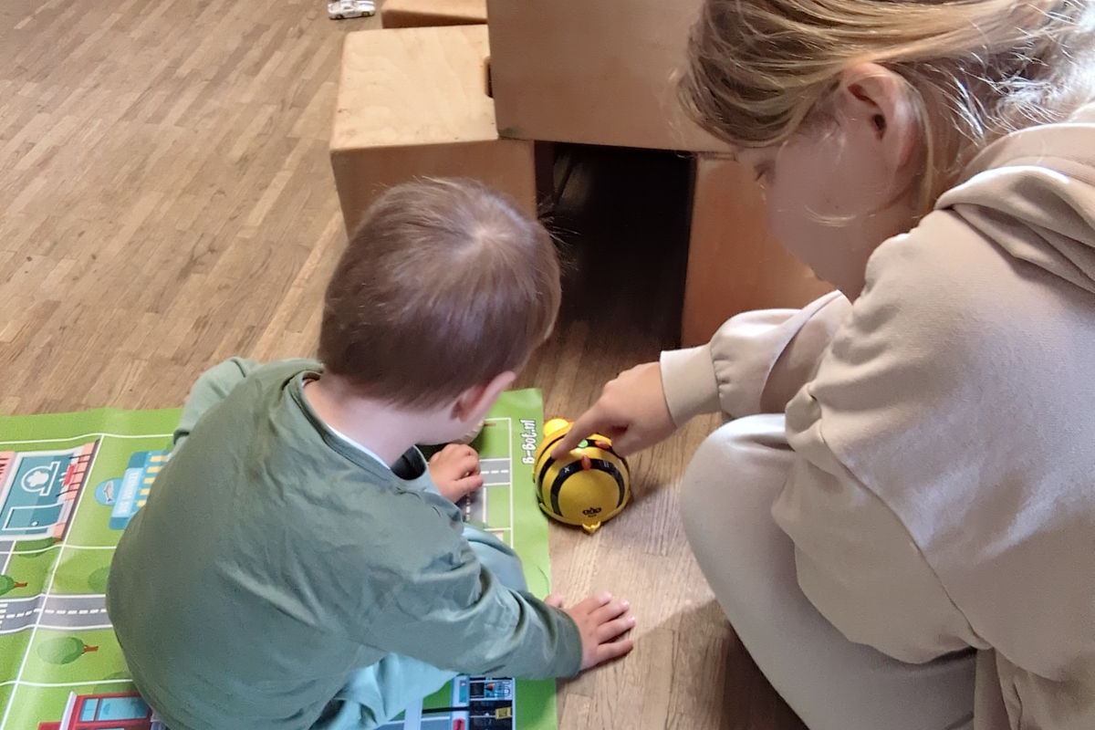 Eine junge Praktikantin und ein Kind spielen mit einer elektronischen Biene. Sie sind von hinten zu sehen.